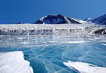 Photograph of blue ice on water in the foreground, with a snowy mountain in the background