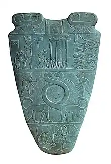 Reverse of the Narmer Palette, circa 3100 BC.  The top row depicts four men carrying standards.  Directly above them is a serekh containing the name of the king, Narmer.