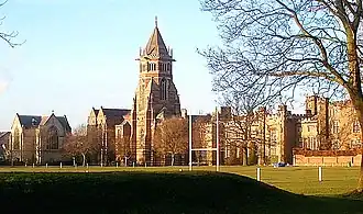 A wide shot of an old English school with a central tower, with a sports pitch in the foreground.