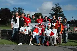 A group of thirteen supporters pose together, nine standing in back row, four seated at front, some wearing rugby jerseys and others sporting traditional Japanese costumes and Japanese flags.